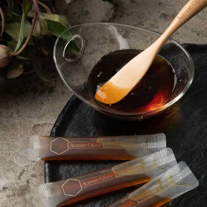 Manuka Honey MGO 150+ in a dish with a wooden spoon and single-serve sachets on a stone surface.