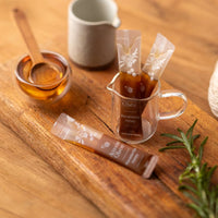 Macadamia Honey sachets in a glass container with a honey dipper and a decorative wooden board.